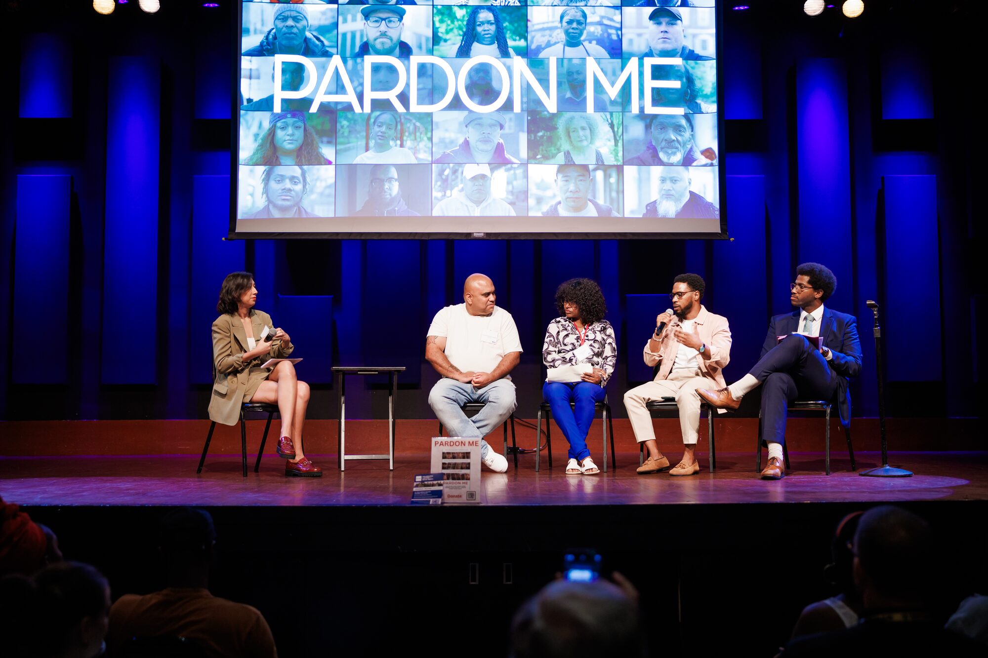 A panel discussion on stage in front of the Pardon Me title screen